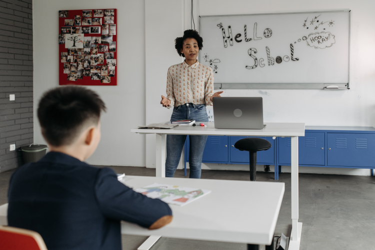Imagem mostra uma professora falando com os alunos em frente a um quadro branco.