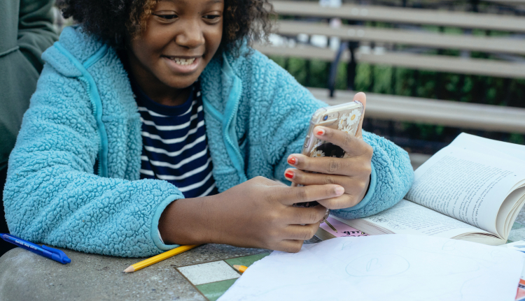 Imagem de uma menina negra bem sorridente, que está com um casaco azul, sentada e segurando e olhando para um celular com as duas mãos.