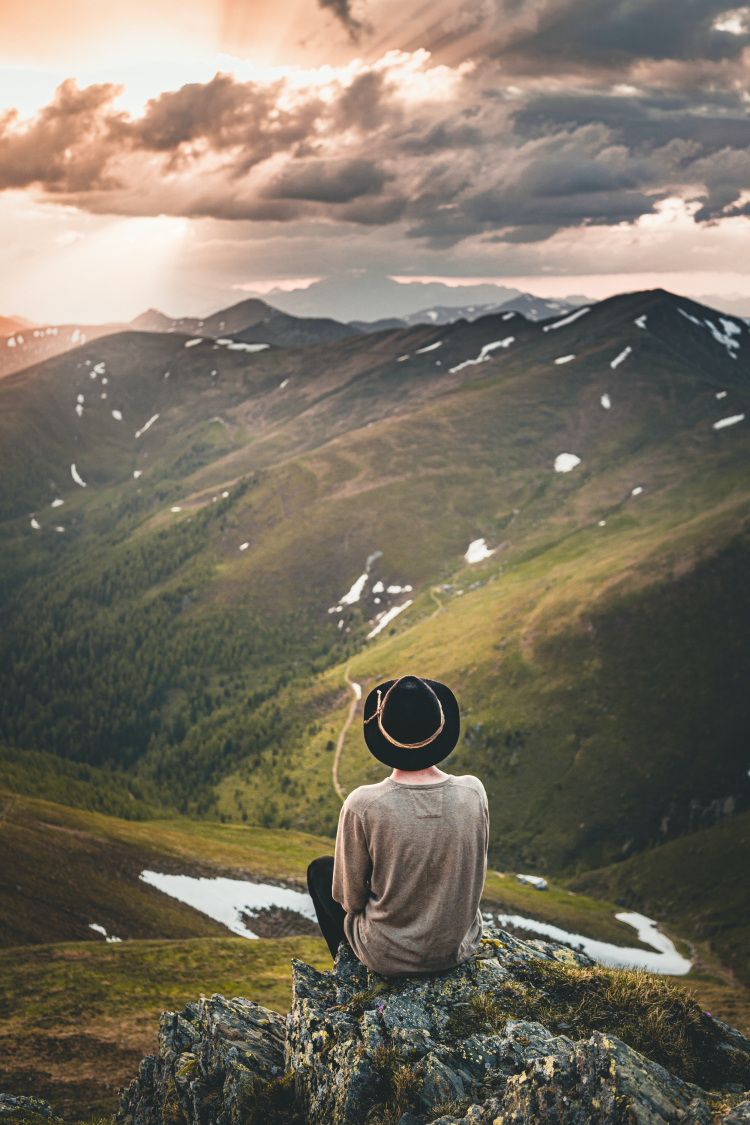 Um homem está sentado em uma rocha admirando a belíssima paisagem à sua frente, que é a de montanhas cobertas por vegetação ao ar livre. Ele está de costas para quem bateu a foto