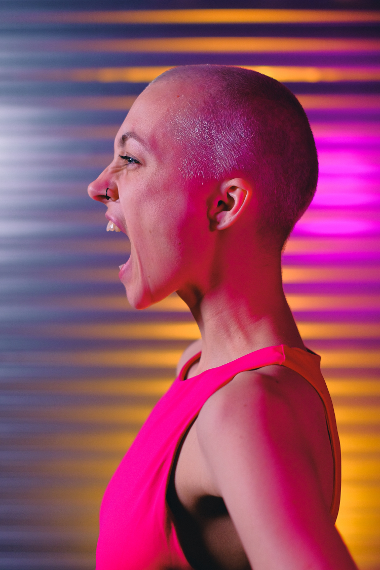 Uma mulher careca está de perfil para quem bateu a foto. Ela está de regata cor de rosa e parece estar gritando