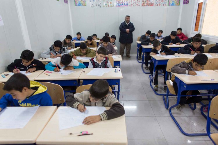 Uma sala de aula com diversos estudantes fazendo uma tarefa enquanto um professor os observa.