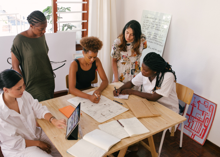 Cionco mulheres estão ao redor de uma mesa, observando e anotando coisas em papeis com informações. Parece uma reunião de trabalho.