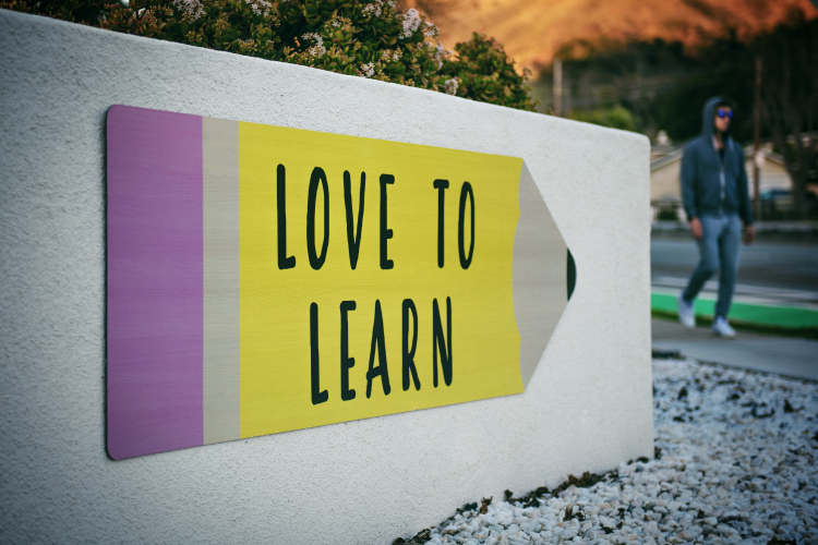 Uma placa no formato de lápis e nas cores amarela e roxa está colada em um muro branco e tem a seguinte frase na parte amarela: "Love to learn"