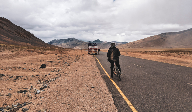 Um ciclista está em pé ao lado de sua bike em uma estrada muito comprida e rodeada por rochas e solo arenoso. Mais à frente na estrada há um furgão ou caminhão estacionado com uma pessoa ao lado