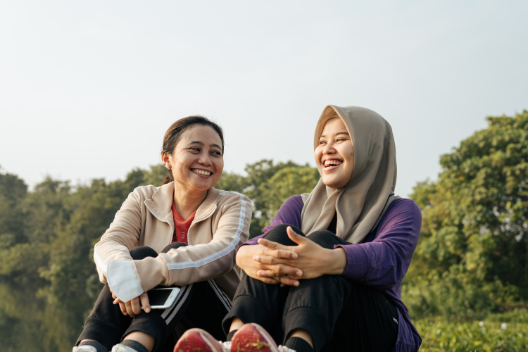 Duas mulheres asiáticas estão sentadas em um campo aberto com muitas árvores ao fundo. Elas estão sorridentes e parecem estar conversando.