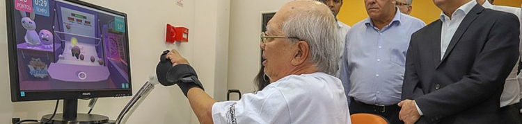 Um idoso branco está com um acessório robótico no braço esquerdo enquanto é observado por outros homens trajando paletó ao lado