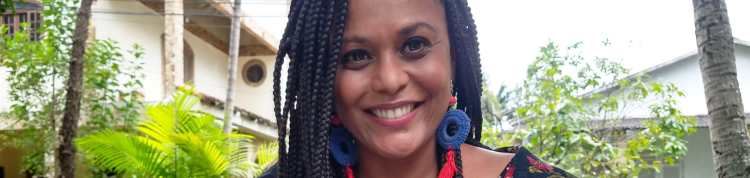 Mulher negra sorri. Ao fundo, o céu e algumas plantas.