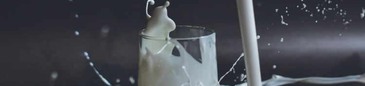 Um copo de vidro está com leite em seu interior e há mais leite sendo derramado ao lado dele, caindo sobre a mesa