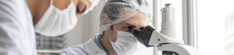 Cientistas de jaleco, máscara de proteção contra covid, luvas e toucas protetoras estão em um laboratório. Um deles está olhando algo em um microscópio