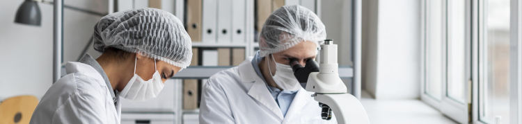 Cientistas de jaleco, máscara de proteção contra covid, luvas e toucas protetoras estão em um laboratório. Um deles está olhando algo em um microscópio