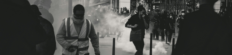 Imagem em preto e branco de uma manifestação urbana que teve confusão, pois há pessoas tapando o rosto com camisas, muita fumaceira, gente correndo, etc