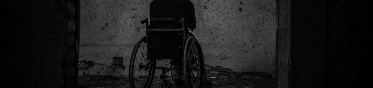 Uma pessoa está sentada em uma cadeira de rodas, de costas, em um local escuro, com pouca iluminação e distante de quem bateu a foto