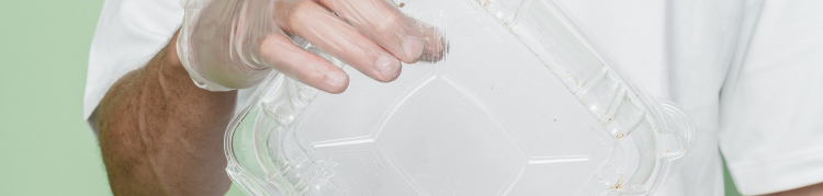 Uma pessoa usando luva descartável está jogando um recipiente de plástico em uma lixeira com a palavra "plastic" escrita