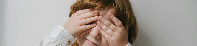 Uma criança branca de cabelos lisos e castanhos está tapando os dois olhos com as suas próprias mãos
