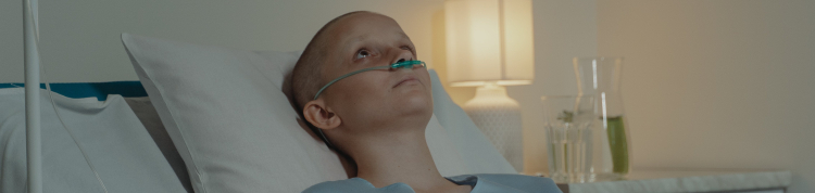 Uma mulher jovem está deitada em uma cama de hospital com tubos de oxigênio. A imagem é em preto e branco e a pessoa está careca, o que nos passa a ideia de ser uma paciente com câncer