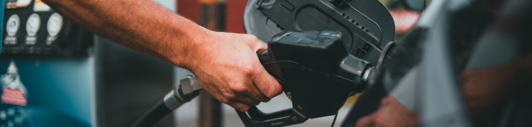Imagem em zoom da mão de uma pessoa branca colocando uma mangueira de gasolina no tanque de um automóvel estacionado em um posto de gasolina