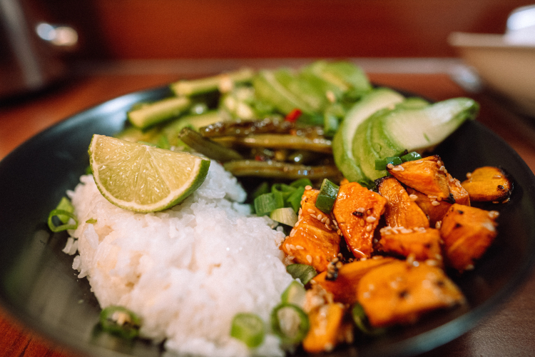 Um prato preto contendo alimentos vegetais e veganos como arroz, batata, limão, picles, vagem