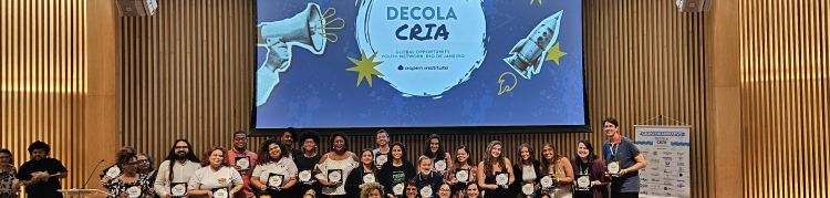 Diversas pessoas (de pé e agachadas, em duas fileiras) sobre um palco. Ao fundo, projetado em tom azulado, o texto "Decola Cria".