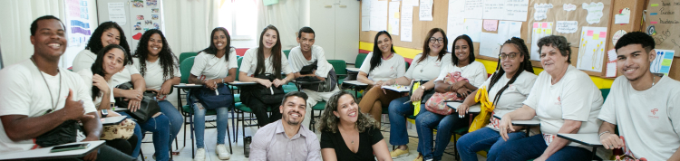 Grupo de estudantes em semi-círculo, sentados em uma sala de aula. No chão, ao centro, há duas pessoas sentadas, um homem e uma mulher.