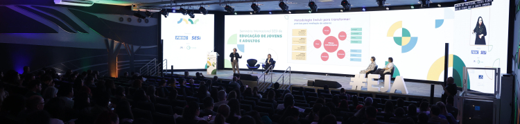 Auditório moderno com cadeiras ocupadas por diversas pessoas assistindo a uma apresentação. No palco iluminado, há quatro pessoas — duas em pé e duas sentadas — participando de um painel com telão ao fundo. No telão está escrito: “Seminário Internacional SESI de Educação de Jovens e Adultos”, com gráficos e textos explicativos. À direita, uma tela vertical mostra uma intérprete de Libras (Língua Brasileira de Sinais). O ambiente tem iluminação colorida nas laterais e teto com painéis de LED.