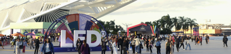 Vista ampla da área externa do Museu do Amanhã, no Rio de Janeiro, durante o Festival LED – Luz na Educação. Em primeiro plano, pessoas caminham e interagem ao redor de uma grande instalação colorida com as letras “LED”. Ao fundo, o mar, palmeiras e tendas coloridas compõem o cenário festivo.