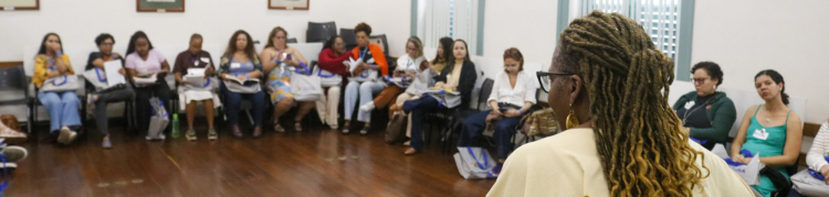 Um grupo de pessoas está sentado em círculo em uma sala. Uma mulher de pé, no centro, fala com o grupo e sorri. A sala tem piso de madeira e quadros retangulares nas paredes.
