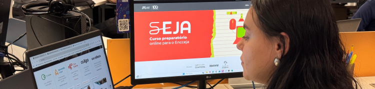 Uma mulher de cabelos longos e escuros, usando brinco de prata, está sentada em frente a duas telas em um ambiente de escritório. Ela olha para um monitor onde está aberto o site do curso “SEJA – Curso preparatório online para o Encceja”. À sua esquerda, há um notebook com a página inicial da Plataforma Futura aberta, exibindo programas como Co.liga, Telê Curso e Aprendê Legal. No fundo, outras pessoas também estão estudando. O ambiente é moderno, com iluminação clara e clima de concentração.