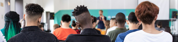 A imagem mostra um grupo de jovens sentados em cadeiras enfileiradas, assistindo a uma apresentação em um ambiente interno, semelhante a uma sala de aula ou auditório. A foto é tirada de trás, mostrando principalmente as costas das pessoas. Três jovens na fileira da frente se destacam: dois com jaquetas pretas e cortes de cabelo estilizados, e um com camiseta branca e cabelo castanho-avermelhado. Ao fundo, há um homem em pé segurando um microfone, aparentemente falando ao público. Atrás dele, há uma tela de