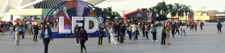 A imagem mostra uma praça com diversas pessoas transitando e um edíficio branco ao fundo, que é o Museu do Amanhã.