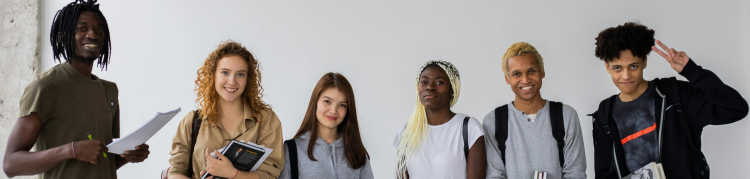 Seis jovens estão alinhados lado a lado, posando para a foto em um ambiente interno de paredes claras. Todos carregam livros ou cadernos e vestem roupas casuais. Da esquerda para a direita: um rapaz de camiseta verde-oliva segura folhas e um lápis; uma jovem de cabelos cacheados ruivos veste camisa bege e sorri; outra jovem de cabelos lisos castanhos usa moletom cinza e segura livros; ao centro, uma jovem de camiseta branca e tranças loiras segura um caderno preto; um rapaz de moletom cinza claro e cabelos 