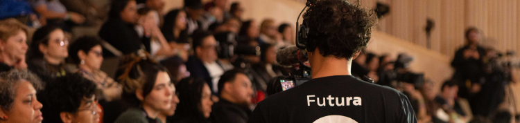 Plateia de um auditório lotado, com pessoas sentadas e atentas à programação. Em primeiro plano, de costas, está um cinegrafista usando fones de ouvido e camiseta preta com o logotipo do Canal Futura, que grava o evento. Algumas pessoas do público seguram celulares e objetos coloridos nas mãos. O ambiente tem iluminação suave e paredes revestidas de madeira clara.