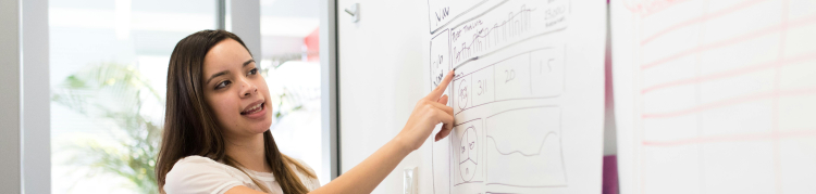 A imagem mostra uma professora explicando algo apontando para um quadro branco. 