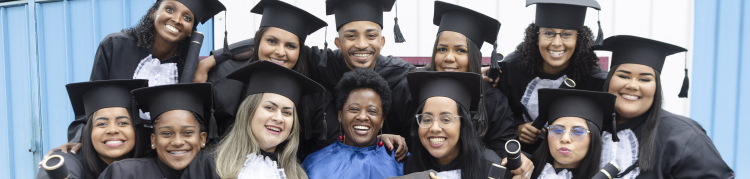 Um grupo diverso de formandos posa sorridente para a foto, usando becas e segurando diplomas. No centro, uma professora também sorri enquanto os abraça. Duas formandas à frente exibem uma placa com a frase: “Melhores professores? Temos.” O clima é de alegria, união e celebração pela conquista.