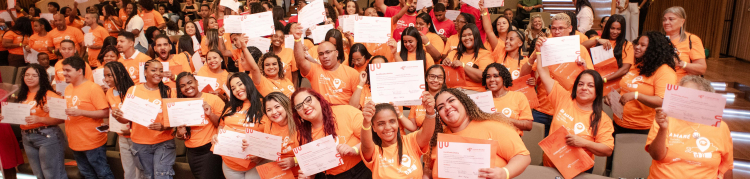 Participantes de uma cerimônia de certificação posam reunidos no auditório, segurando seus certificados com entusiasmo. Os formandos, vestidos com camisetas laranja, ocupam a frente da sala, enquanto o público acompanha a celebração nas arquibancadas ao fundo. A cena transmite sentimento de orgulho coletivo e comemoração.