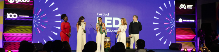 Em um palco de evento, cinco pessoas estão em pé, alinhadas, diante de uma plateia numerosa sentada. O público aparece em primeiro plano, visto de costas e em silhueta. No centro do palco, uma mulher fala ao microfone; à sua frente há dois tambores altos, decorados com padrões coloridos.  O cenário ao fundo é um grande painel roxo com a inscrição “Festival LED Educação”, acompanhado de elementos gráficos que lembram raios ou explosões coloridas nas laterais. 