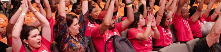 Auditório cheio de pessoas sentadas em poltronas, participando de um evento coletivo. Em primeiro plano, várias pessoas vestem camisetas vermelhas e levantam os braços acima da cabeça, aplaudindo ou comemorando juntas. As expressões são de entusiasmo e envolvimento. Algumas pessoas usam pulseiras e crachás. Ao fundo, o auditório segue lotado, com público em diferentes níveis de assentos, todos voltados para a frente do espaço.