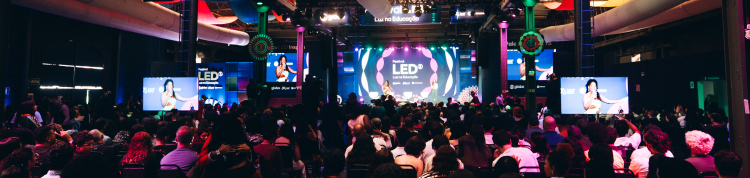 Auditório amplo e escuro, iluminado por luzes coloridas em tons de rosa, azul e roxo. Centenas de pessoas estão sentadas em cadeiras voltadas para um grande palco central, onde acontece uma apresentação do Festival LED – Luz na Educação. No fundo do palco há um telão circular com a identidade visual do evento, cercado por painéis luminosos e dois telões laterais exibindo a palestrante. O teto industrial alto possui estruturas metálicas aparentes e grandes dutos brancos atravessando o espaço